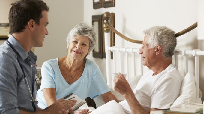 Doctor Having A Conversation At The Bedside Of An Elderly Man And His Wife.