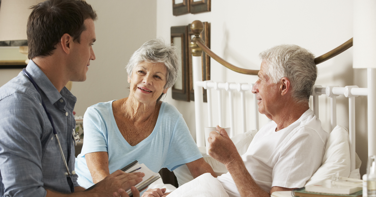 Doctor Having A Conversation At The Bedside Of An Elderly Man And His Wife.