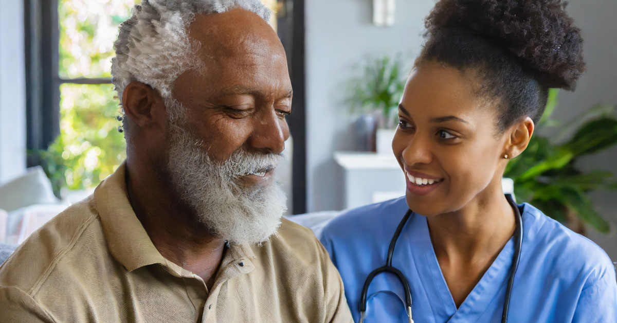 African American Nurse Lifting The Spirits Of An Elderly Man In Hospice.